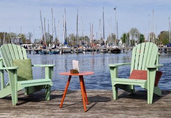Two green chairs and a table on a wooden dock by the water with a view of a marina on a sunny day.