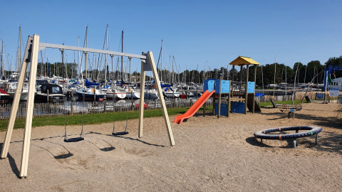 Spielplatz mit Schaukeln und Rutsche am Hafen voller Segelboote, aufgenommen bei klarem Himmel.