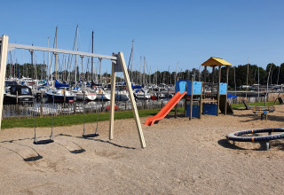 Spielplatz mit Schaukeln und Rutsche am Hafen voller Segelboote, aufgenommen bei klarem Himmel.