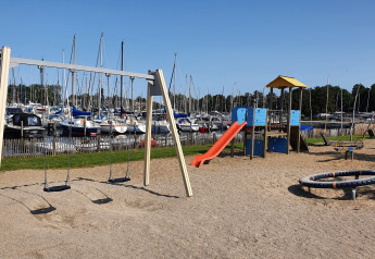 Aire de jeux avec balançoires et toboggan près d’un port de voiliers, sous un ciel bleu dégagé.