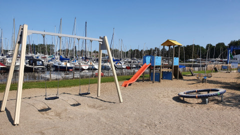 Legeplads med gynger og rutsjebane ved havn fyldt med sejlbåde, fotograferet under blå himmel.