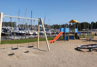 Legeplads med gynger og rutsjebane ved havn fyldt med sejlbåde, fotograferet under blå himmel.