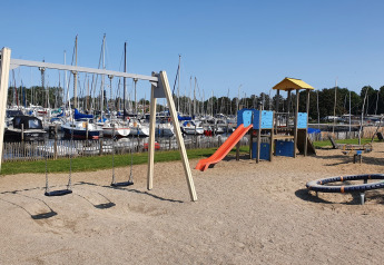 Legeplads med gynger og rutsjebane ved havn fyldt med sejlbåde, fotograferet under blå himmel.