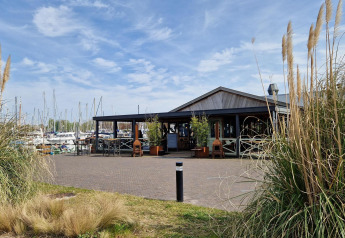 A cozy houseboat café area by the marina with boats in the background and tall reeds in the foreground.