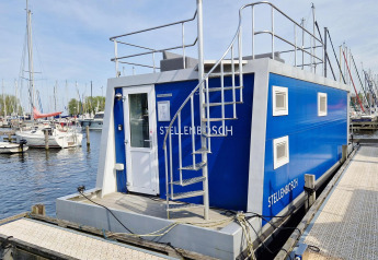 Blå husbåd ved navnet Havenlodge N fortøjet ved Marina Parcs Naarden i Holland, omgivet af sejlbåde.