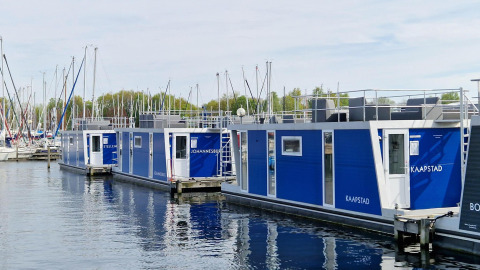 Modernas casas flotantes con paneles azules atracadas en un puerto deportivo, con veleros al fondo.
