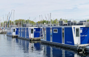 Modernas casas flotantes con paneles azules atracadas en un puerto deportivo, con veleros al fondo.