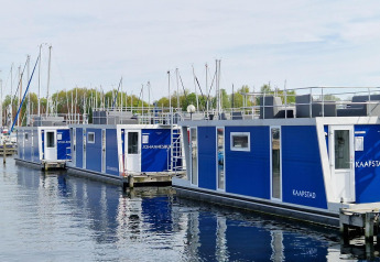 Modern houseboats with blue panels docked at a marina, sailboats and trees visible in the background.