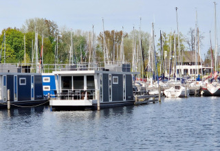 Casa flotante llamada Havenlodge N en Marina Parcs Naarden, Países Bajos, junto a veleros y árboles verdes.