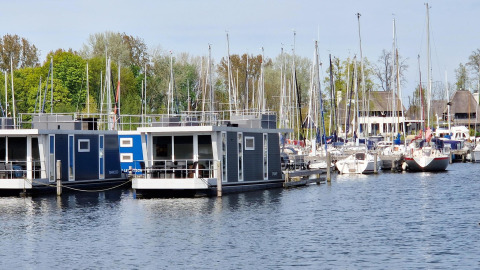 Casa flotante llamada Havenlodge N en Marina Parcs Naarden, Países Bajos, junto a veleros y árboles verdes.