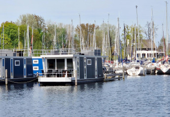 Woonboot Havenlodge N in Marina Parcs Naarden, Nederland, aangemeerd tussen zeilboten en bomen.