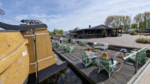 Folk nyder solskin ved vandet på en glampingplads med stole, en båd og restaurant i baggrunden.