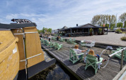 Folk nyder solskin ved vandet på en glampingplads med stole, en båd og restaurant i baggrunden.