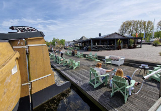 Folk nyder solskin ved vandet på en glampingplads med stole, en båd og restaurant i baggrunden.