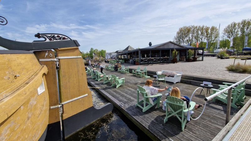 Personas descansan junto al agua en sillas de madera en un alojamiento glamping, con bote y restaurante cerca.