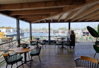 Zicht vanaf een overdekt terras met cafetafels op een jachthaven bij een glampingverblijf.