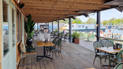Überdachte Terrasse auf einem Hausboot mit Tischen, Stühlen und Blick aufs Wasser und Boote.