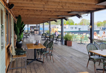 Udendørs terrasse på en husbåd med borde, stole og udsigt over vandet og både i baggrunden.