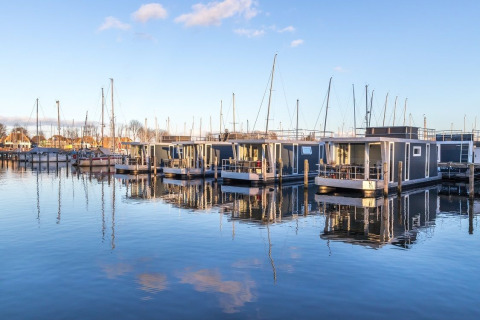 Moderne woonboten liggen aangemeerd in een rustige haven met helder water en een blauwe lucht op de achtergrond.