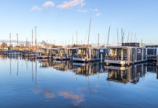Moderne woonboten liggen aangemeerd in een rustige haven met helder water en een blauwe lucht op de achtergrond.