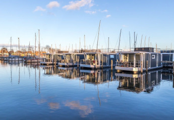 Moderne husbåde ligger fortøjet i en rolig havn med klart vand og en blå himmel i baggrunden.
