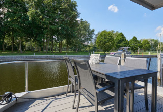 Udsigt fra husbådens terrasse med bord og stole ved vandet, grønne træer og både i baggrunden.