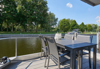 View from houseboat deck with outdoor table and chairs by the water, green trees and boats in background.