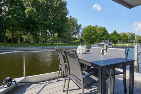 View from houseboat deck with outdoor table and chairs by the water, green trees and boats in background.