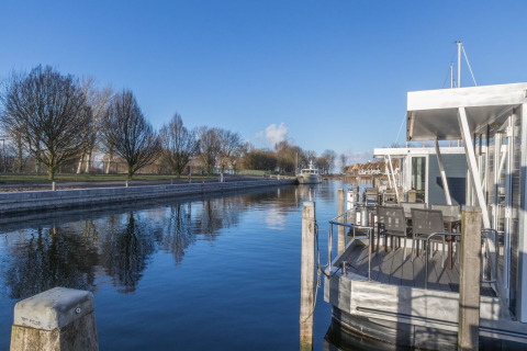 Foto van een woonboot aangemeerd langs een rustige waterweg, bomen en blauwe lucht op de achtergrond.
