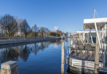 Foto af en husbåd fortøjet ved en rolig kanal med træer og blå himmel i baggrunden, stille atmosfære.