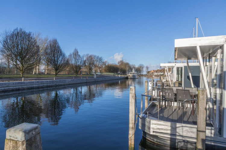 Foto de una casa flotante atracada junto a un canal tranquilo con árboles y cielo azul al fondo.