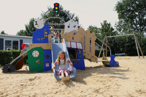 Børn leger på legeplads med rutsjebane og gynger i sandet ved Sunlodge på Holiday Park 't Rheezerwold.