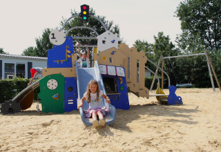 Børn leger på legeplads med rutsjebane og gynger i sandet ved Sunlodge på Holiday Park 't Rheezerwold.