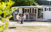 Niños juegan delante de un chalet familiar mientras los padres descansan en la terraza en Kampeerdorp de Zandstuve.