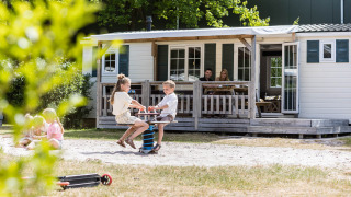 Bambini giocano davanti a uno chalet familiare mentre i genitori si rilassano sul portico a Kampeerdorp de Zandstuve.