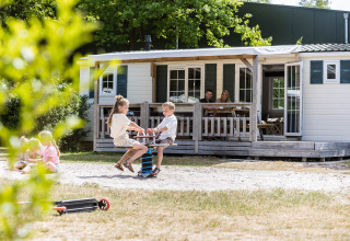 Børn leger foran en familiechalet med forældre, der slapper af på verandaen i Kampeerdorp de Zandstuve.