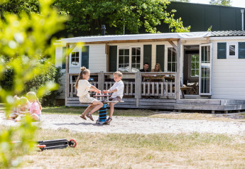 Kinder spielen vor einem Familienchalet, während Eltern auf der Veranda bei Kampeerdorp de Zandstuve entspannen.