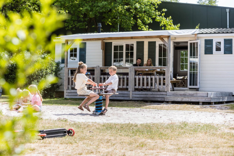 Niños juegan delante de un chalet familiar mientras los padres descansan en la terraza en Kampeerdorp de Zandstuve.