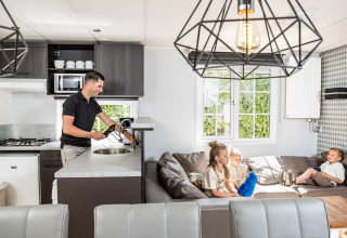 Man washing dishes in the kitchen while three children play on the sofa in a family chalet at Kampeerdorp de Zandstuve.