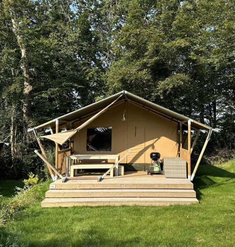 Tenda safari con terrazza in legno, panca e barbecue, immersa nel verde tra erba e alberi alti.