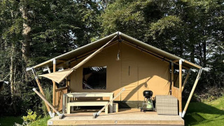 Safari tent with a wooden deck, seating, and grill set among lush trees and grass in a natural setting.