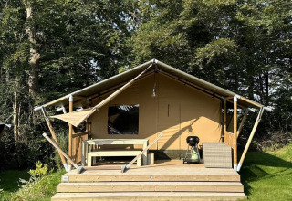 Safari telt i naturen med træterrasse, udendørs bænk og grill foran, omgivet af grønne træer og græs.