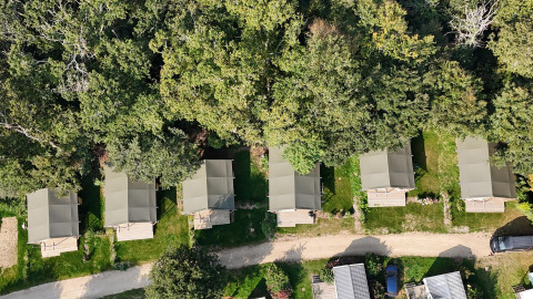 Aerial view of six safari tents lined up by the forest edge at a green campsite with a dirt path.
