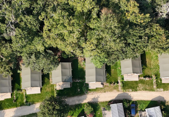 Aerial view of six safari tents lined up by the forest edge at a green campsite with a dirt path.