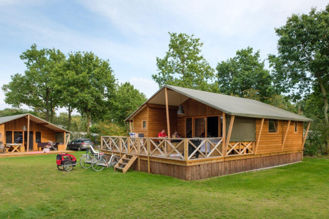 Außenaufnahme von Rheezerlodge DeLuxe high im Kampeerdorp de Zandstuve, Niederlande, mit Fahrrädern und Hütten.
