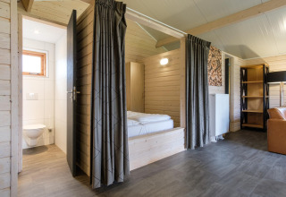 Moderne lodge med lyst trægulv, synlige bjælker, soveværelse med gardiner og tilstødende badeværelse.