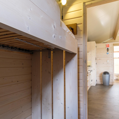 Interior image of a lodge with light wood panel walls, bunk bed, and view into a bathroom with a bin.