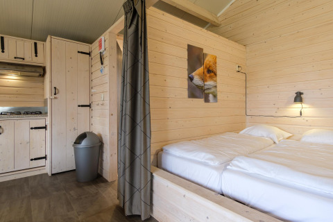 Interior de una acogedora cabaña con paredes de madera clara, cama doble, cocina pequeña y cuadro de zorro.