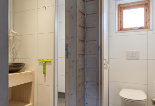 Moderne lodgebadkamer met witte tegels, wastafel, haardroger en apart toilet met houten raam.