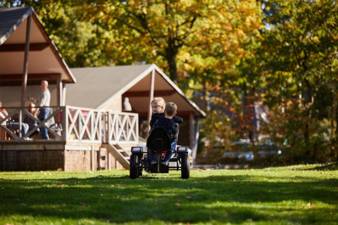 Two children ride a go-kart on a grassy area in front of cabins at a scenic, wooded lodge.